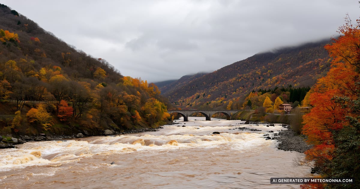 Un fiume appenninico gonfio di acqua scura tra colline boscose in autunno, con nuvole basse e grigie sulle creste e un ponte in pietra sullo sfondo.